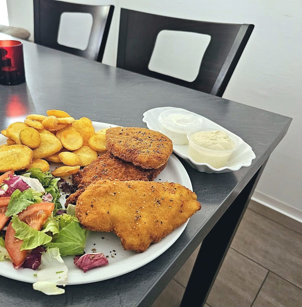 Drei panierte Schnitzel mit Kartoffelscheiben, gemischtem Salat und zwei Dips auf einem Teller